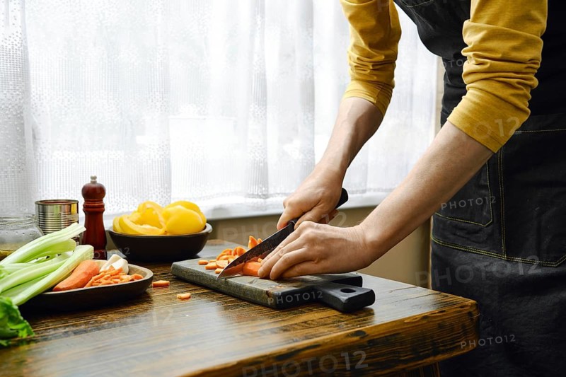 In a kitchen with natural light, a person is chopping carrots on a wooden cutting board. Fresh vegetables are on the table, including celery and lemon, as they prepare a meal