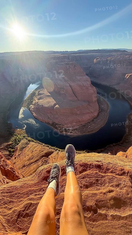 Adventurous traveler dangling legs over the edge of horseshoe bend canyon, showcasing the vast expanse of the colorado river and sandstone rock formations under a bright sky