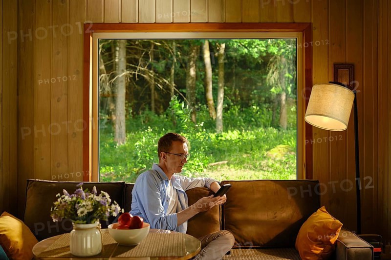 Middle-aged man sits on couch in wooden cabin, holding electronic book and reading a novel. Light streams through large window. Green trees show outside