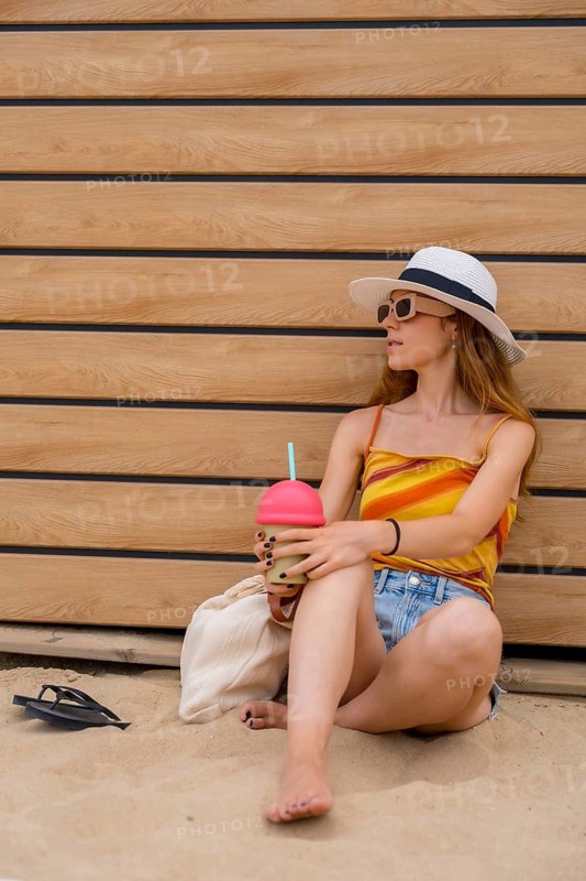 Young woman relaxing barefoot on the sand, dressed in summer clothes, wearing a hat and sunglasses, holding a refreshing drink while leaning against a wooden wall, enjoying her seaside vacation