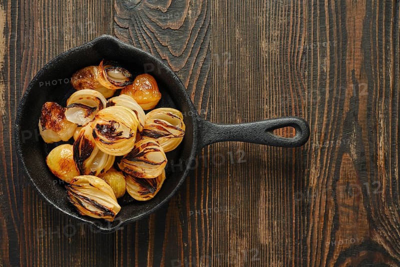 Top view of cast iron skillet with roasted onions on wooden table. Onions show grill marks and slight caramelization. Natural light highlights their texture and color