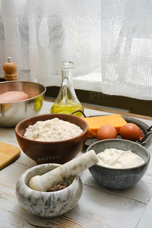 Different cooking ingredients and tools are placed on a table near a window. There is flour in a bowl, eggs, cheese, oil, and a mortar and pestle. This scene shows a cooking preparation area