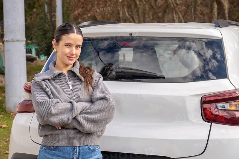 Confident young woman standing with arms crossed beside rear of white car, outdoor rural setting, new driver portrait concept