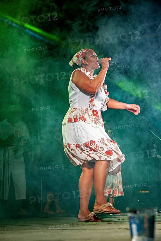 Woman singing on stage in a white dress with red floral patterns in the fog in front of colorful lights, a band plays and dances at a festival in Petrolina in Brazil
