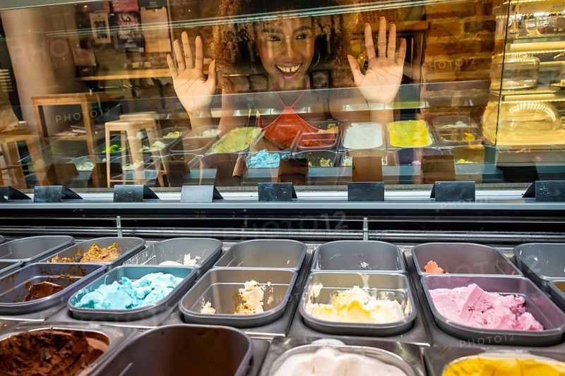 Young woman with curly hair pressing hands against a glass freezer door, smiling and looking at various vibrant flavors of tempting gelato and ice cream in a shop