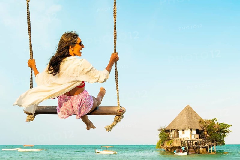 Carefree excited Middle aged woman tourist in pink sarong relaxes on beach swing overlooking the iconic Rock Restaurant in Zanzibar. Peaceful coastal scene with turquoise water. Travel holidays vacation