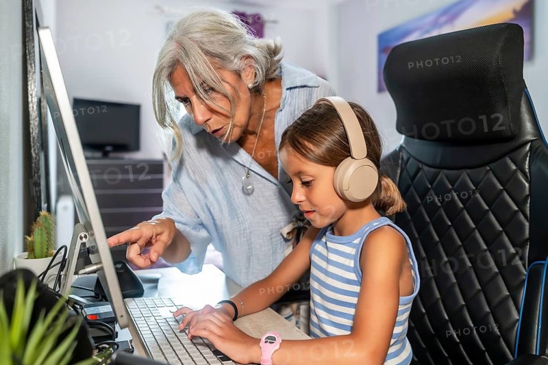 Grandmother is patiently teaching her young granddaughter how to use a computer. Pointing at the screen and guiding her as the child types on the keyboard