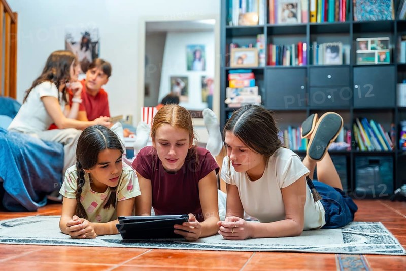Three sisters are lying on the floor looking at a tablet together while two older siblings are talking on a bed in the background, depicting a modern family's casual indoor leisure time