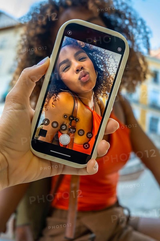 Hand holding a smartphone displaying a smiling young woman making a kissing face for a selfie, capturing a moment of joy and connection outdoors during travel