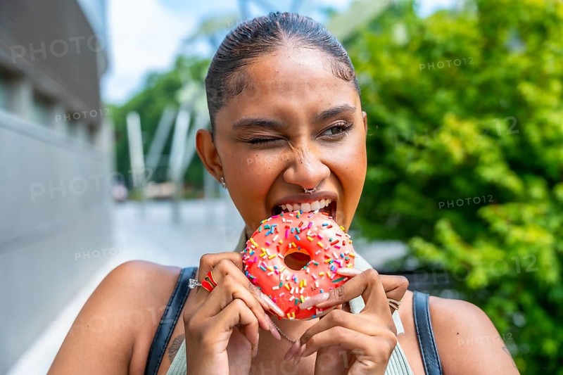 Young woman with a nose ring making a playful face while eagerly biting into a vibrant pink donut with colorful sprinkles, enjoying a sweet treat outdoors on a sunny day