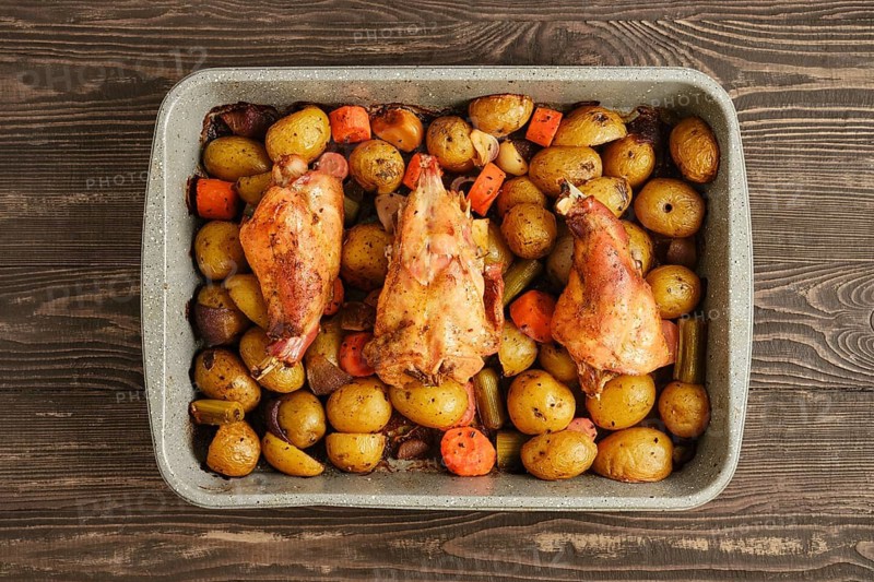 Rabbit meat and assorted vegetables are cooking together in a baking dish. The dish sits on a wooden table, ready for a family meal. The food is warm and inviting for dining