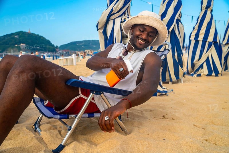 Young man relaxing on a beach chair under striped umbrellas, smiling and applying sunscreen to his arm, embracing sun protection during summer vacation