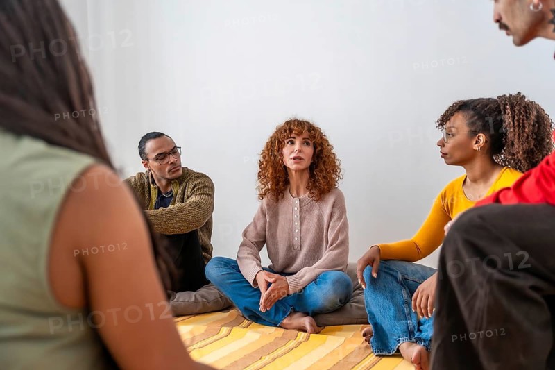 Diverse adults are sitting on floor cushions in a circle, participating in a group therapy session, sharing experiences, listening actively, and receiving supportive mental health counseling
