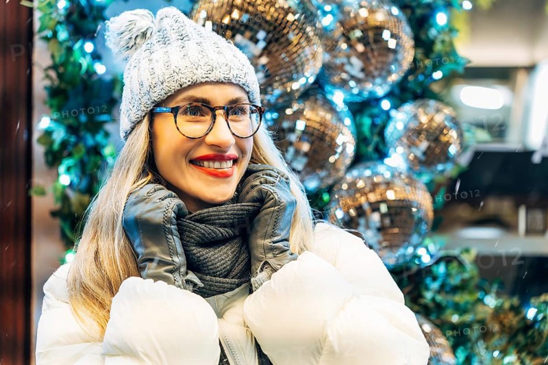 Young woman in winter clothing with festive decorations. A person in a white puffer jacket, gray scarf, knit hat with a pom-pom and glasses stands near large silver disco balls surrounded by greenery and blue-white lights. The reflective surfaces capture surrounding illumination, creating bright highlights and layered reflections. The close framing emphasizes winter clothing textures and the vivid festive lighting around the figure.z