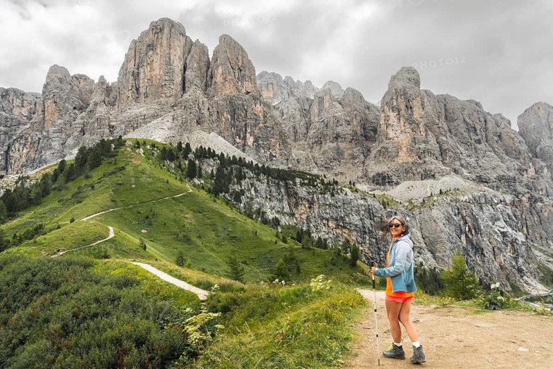 Female hiker with trekking pole turning to look at camera walking winding dirt path along green alpine ridge majestic jagged Cir peaks background Passo Gardena Dolomites Italy