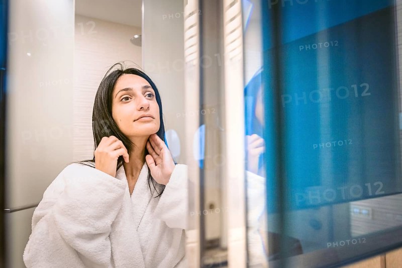 Pretty woamn brushes her hair and looks to her bright clear skin in the mirror with reflection light in the background. Batumi. Georgia