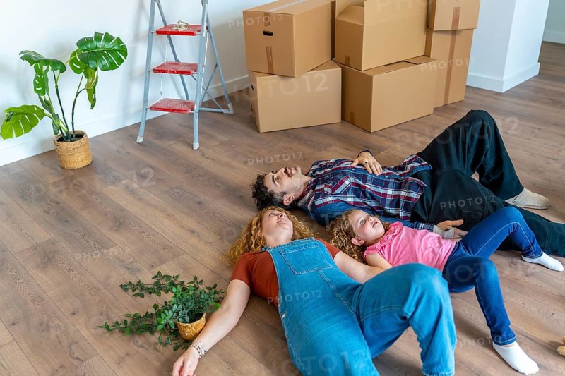 Family parents and daughter resting on the floor in their new home, surrounded by unpacked moving boxes and household plants, pausing after the relocation process