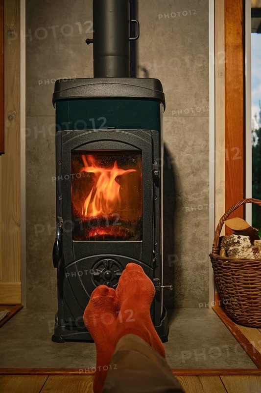 Feet in red socks rest in front of wood stove with flames. Basket nearby shows logs. Room has wooden walls. Light from fire offers simple warmth at night