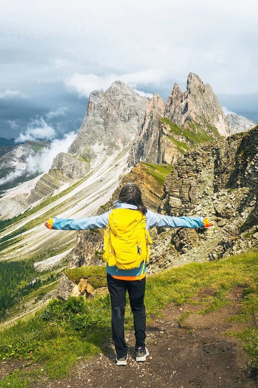 Back view middle aged gray hair woman hiker arms outstretched with yellow backpack overlooking lush green alpine valley misty mountains background Dolomites Alps Italy. Seceda ridge viewpoint