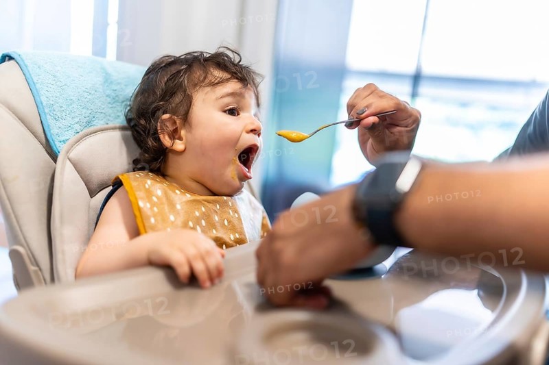 Young baby boy sitting in a high chair opening his mouth wide, being fed healthy vegetable puree by a parent's hand with a spoon, showing a happy mealtime at home