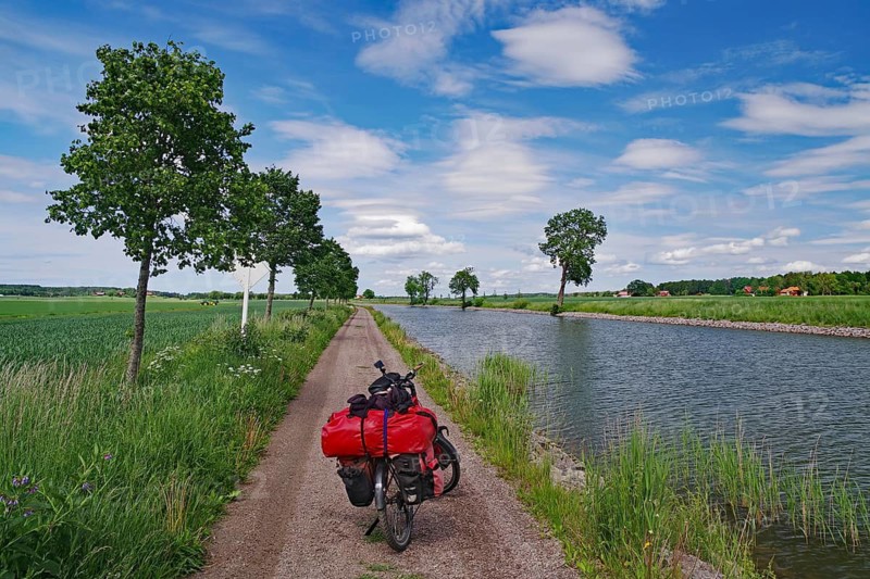 Cycling on a narrow canal path with a landscape of water and trees under a blue sky, bicycle tours, Göta Canal, Töraboda, Västra Götalands län, Sweden