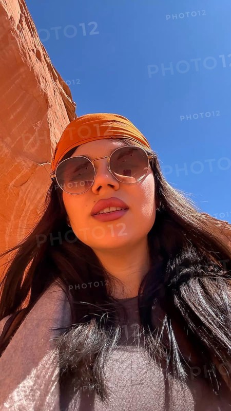 Woman wearing sunglasses and an orange head bandana posing for a close up selfie while exploring the vibrant orange sandstone walls of antelope canyon in arizona under a clear blue sky