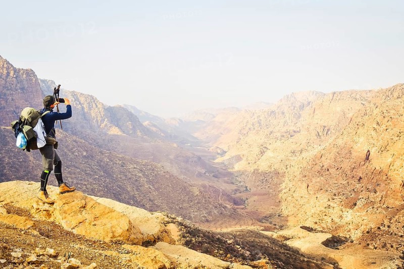 Caucasian Man hiker with large backpack standing rocky ridge taking smartphone picture panoramic sandstone canyon landscape viewpoint hiking travel adventure Dana Reserve Jordan