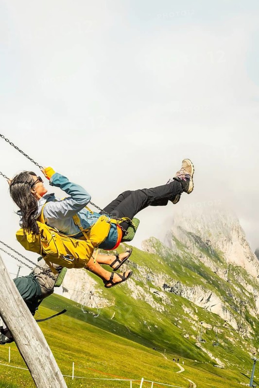 Excited woman with backpack swinging on wooden playground swing lush green hills steep mountain peak misty background Seceda Dolomites Italy summer travel adventure lifestyle