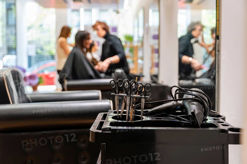 Hairdressing tools including scissors and a hairdryer resting on a trolley in the foreground with a blurred stylist cutting a client's hair in the background, showing a typical working environment