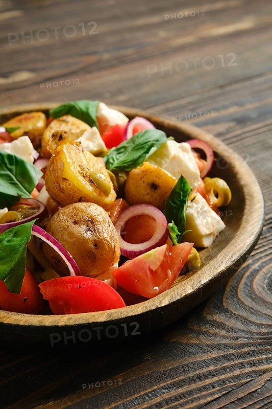 Close-up view of warm potato and feta salad is served in wooden bowl. It includes potatoes, feta cheese, tomatoes, onions, olives, and fresh basil