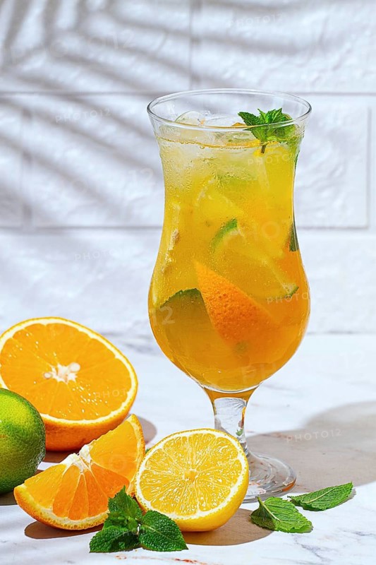 A glass with a drink made from orange and lime sits on a marble table. Fresh orange and lime slices are next to the glass, and there are mint leaves for garnish