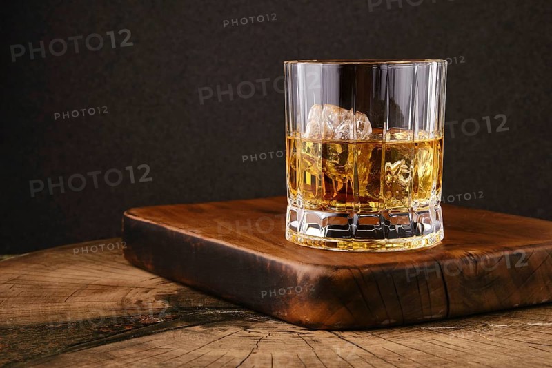 Facetted glass filled with whiskey and ice on wooden board in kitchen. Dark background highlights drink and glass shape. Light reflects off ice showing clarity of drink