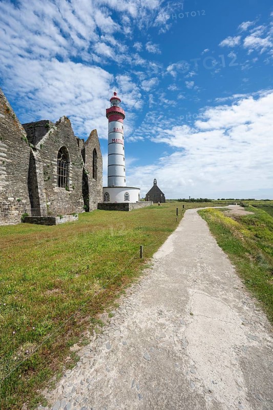 View of the 37 meter high, red-and-white striped lighthouse Phare de Saint-Mathieu, which rises majestically amidst the historic ruins of an old medieval former Benedictine abbey, Abbey Saint-Mathieu de Fine-Terre. A paved footpath leads through a green coastal landscape under blue summer skies with white clouds directly to the historic landmark. Pointe Saint-Mathieu, Plougonvelin, Arrondissement of Brest, Finistère Department, Brittany Region, France