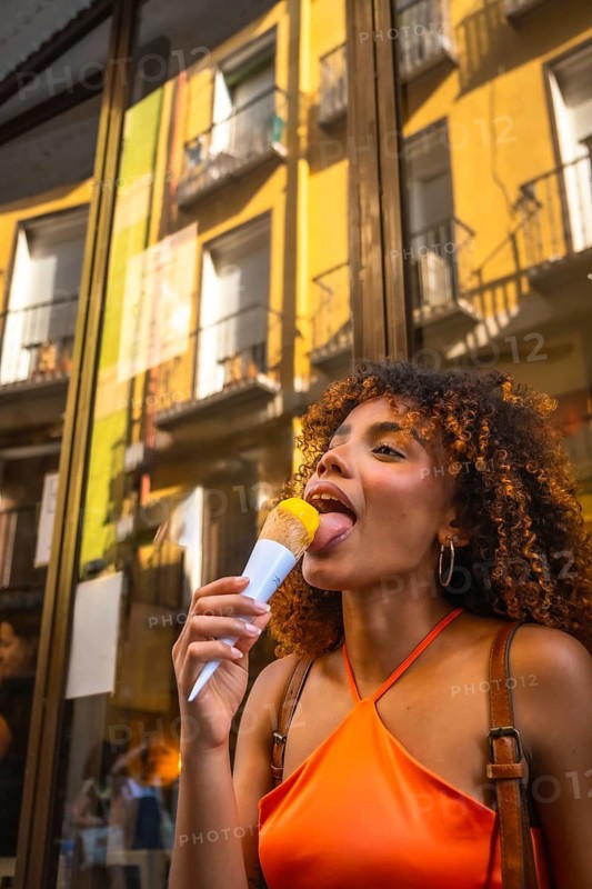 Young woman with curly hair wearing an orange top licking a yellow ice cream cone, experiencing refreshing delight on a sunny street with building reflections