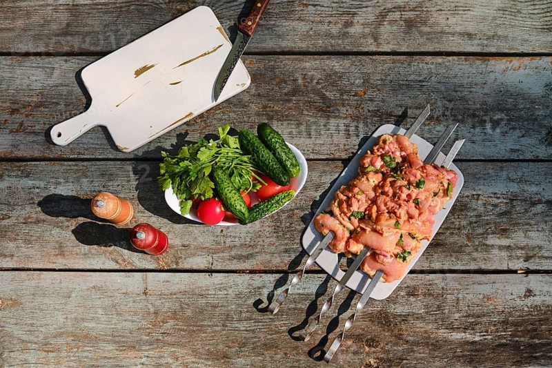 Top view of marinated chicken pieces on skewers beside tomatoes, cucumbers, and herbs on wooden table outdoor