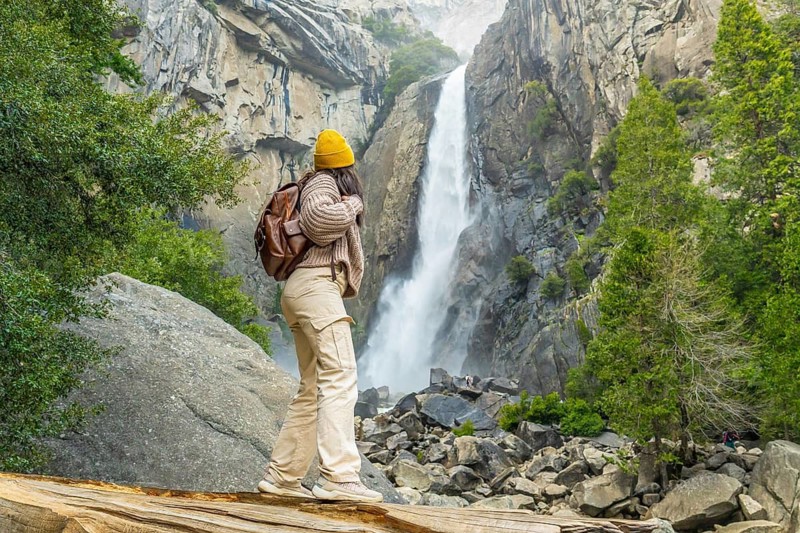 Woman standing on a fallen log, looking at the powerful flow of lower yosemite falls and feeling inspired by the majestic natural beauty of the yosemite national park landscape
