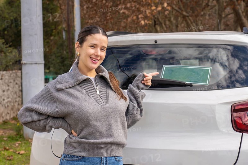 Smiling young woman pointing at blank green sign on rear car window, outdoor rural setting, customizable sign concept