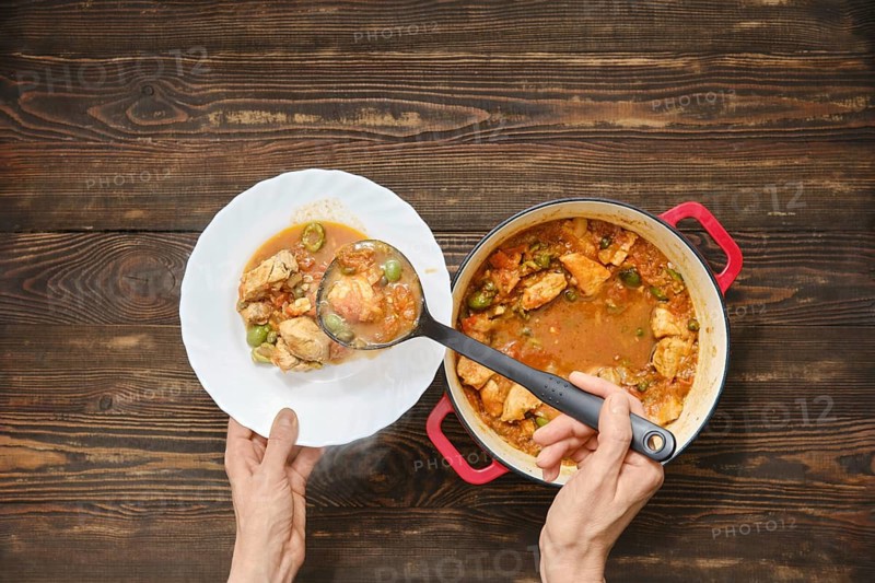 Top view of person using ladle to serve meat stew into white plate. Pot is full of stew with vegetables and rests on wooden tabletop