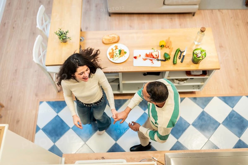 Joyful couple holding hands and dancing together in their home kitchen, sharing a loving and fun moment while preparing fresh vegetables on a wooden island countertop