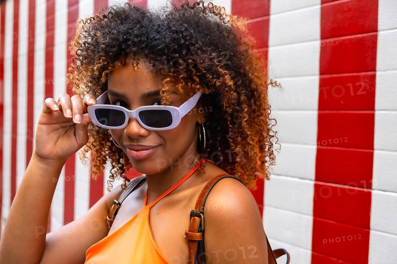 Confident young woman with curly hair pulling down light purple sunglasses and smiling, wearing an orange top against a vibrant red and white striped background