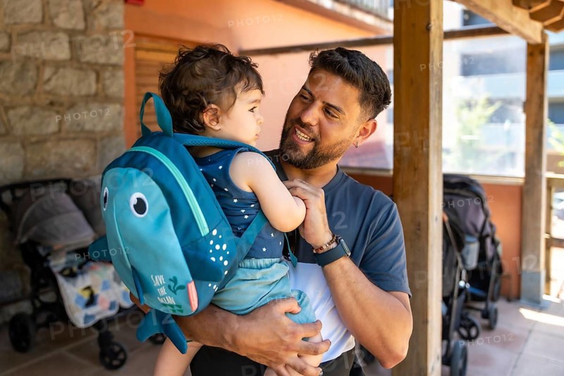 Father holding his young son, who is wearing a fish shaped backpack, smiling and looking at him with affection and support, capturing a moment of family bonding