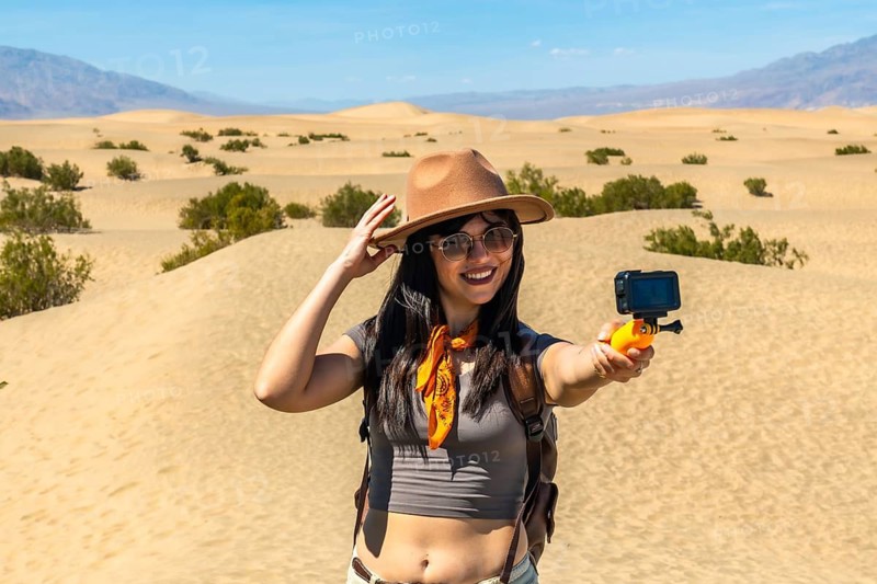 Young woman standing on the mesquite flat sand dunes in death valley national park, holding a camera on a selfie stick and smiling, creating travel video content for social media