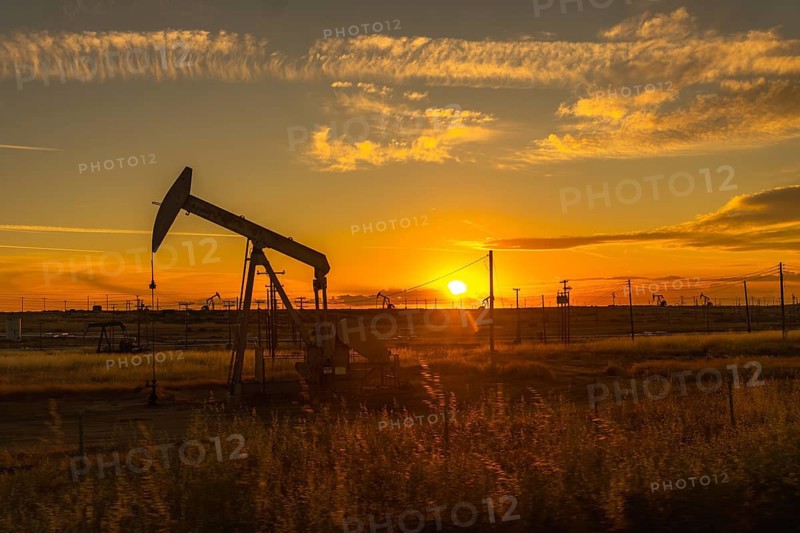 Pumpjack extracting crude oil from a well in a large oil field, silhouetted against a vibrant orange and yellow sunset sky with scattered clouds, representing the energy industry