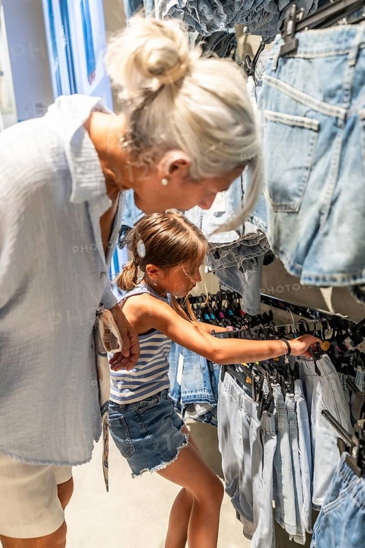 Grandmother assisting her young granddaughter choosing denim shorts from a clothing rack in a high street shop, enjoying a family shopping trip together