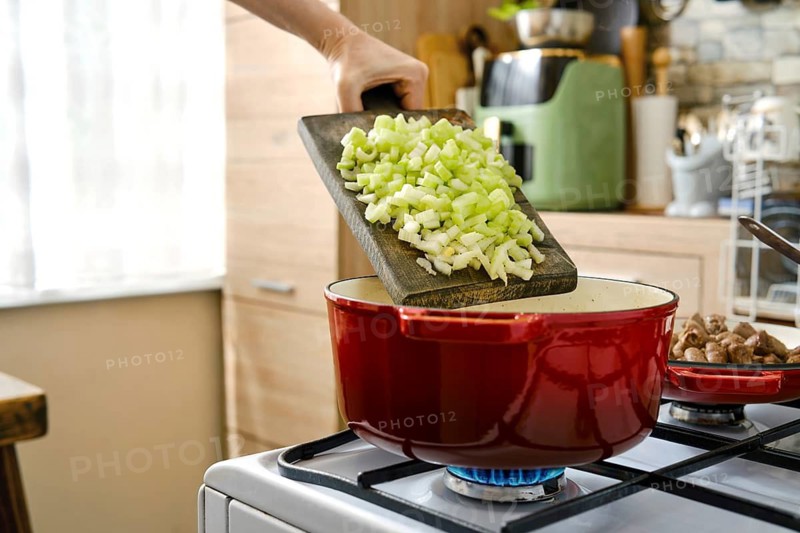 A hand holds a cutting board with chopped celery above a red pot on a stove. The kitchen has natural light and various cooking tools. The person is preparing food in the afternoon