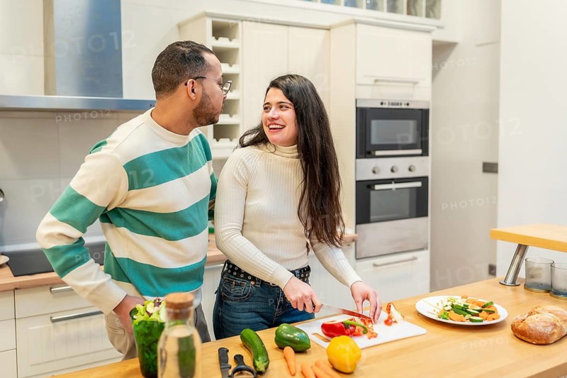 Young happy couple standing in a bright kitchen, the woman preparing fresh vegetables while looking at the man and smiling, sharing a healthy lifestyle moment together