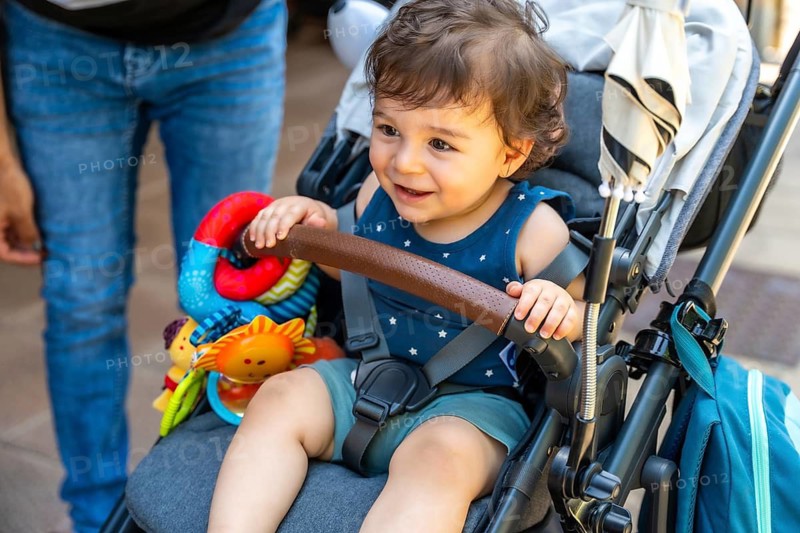 Smiling toddler sitting securely in a modern stroller, holding onto the safety bar and observing the surroundings with a joyful expression while a parent stands nearby, ready for an outdoor activity