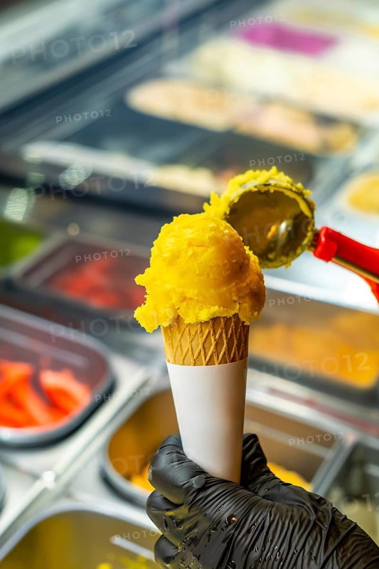Hand in black glove scooping vibrant yellow mango sorbet into a crispy waffle cone from a gelato display case, creating a refreshing summer treat perfect for warm weather