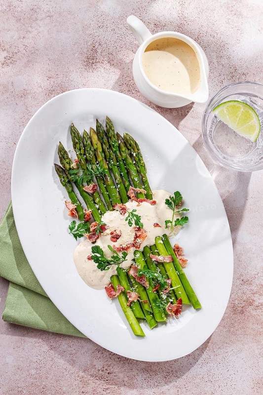 Fresh asparagus with cream sauce and bacon, on a white oval plate, light marble table, sunlight, homemade, no people