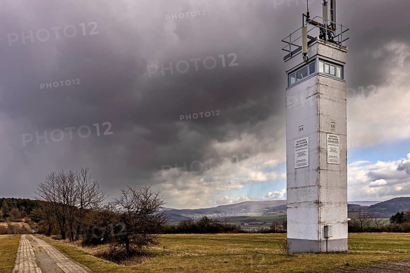 A watchtower stands under a dramatic cloudy sky in a meadow next to a small road, The Point Alpha Memorial between Rasdorf and Geisa in the Rhön